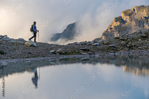 A person stands on rocky terrain, gazing at a stunning turquoise mountain lake surrounded. Dressed in hiking gear and holding trekking poles. Wildsee Pizol, Switzerland.