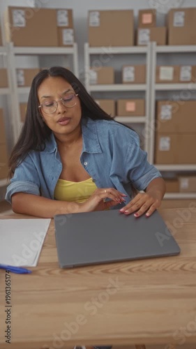 Woman with glasses in denim shirt closes laptop with hand and rubs forehead at a storage building with shelves of boxes; small business fatigue.