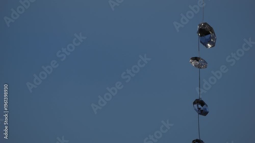 4K telephoto-style shot of decorative glass crystals suspended on a string, gently swaying in the wind against a clean blue sky background. The composition emphasises minimalism, negative space, and s