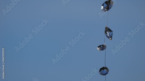 4K telephoto-style shot of decorative glass crystals suspended on a string, gently swaying in the wind against a clean blue sky background. The composition emphasises minimalism, negative space, and s