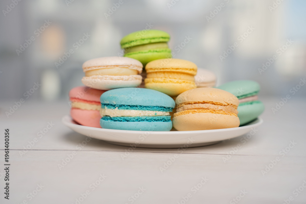 Fototapeta premium close-up of a pastel assortment of colorful french macarons stacked on a white plate on a pale wooden table with soft natural bokeh background, creating a delicate airy dessert scene, shallow
