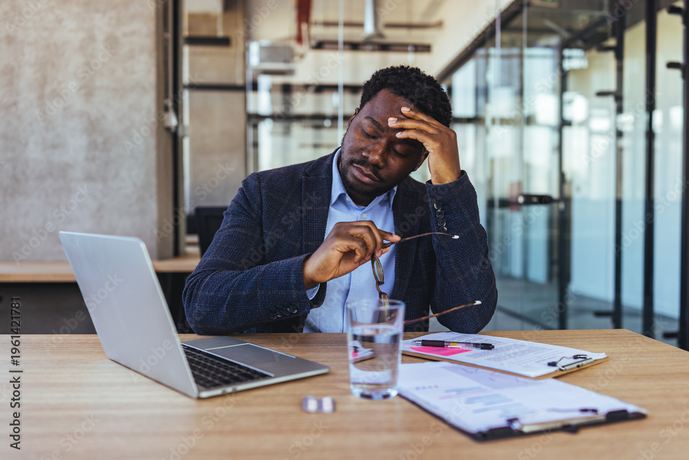 Fototapeta premium Tired Businessman At Office Desk Rubbing Forehead With Glasses Laptop And Documents