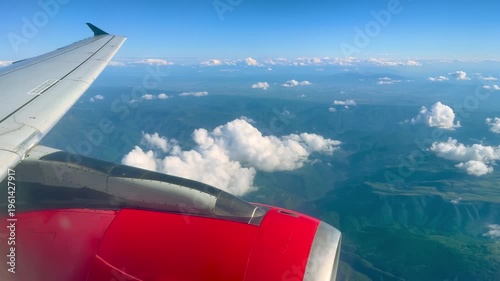View from inside passenger airplane showing red jet engine through window over mountain landscape and clouds during flight in daylight
