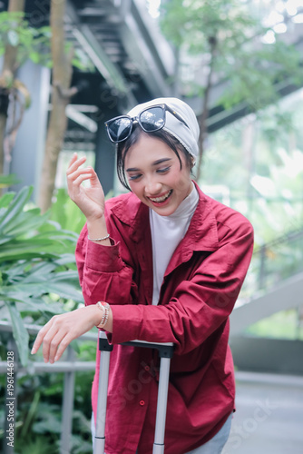 A stylish and happy young woman with a hijab and sunglasses is traveling. She is smiling and looking down while holding her suitcase, radiating happiness and confidence as she embarks on her journey.