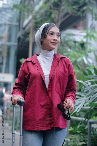 Elegant young woman adorned with a hijab, joyfully strides onward, pulling luggage outside an airport. She exudes an aura of confidence and embraces contemporary fashion while on a global escapade.