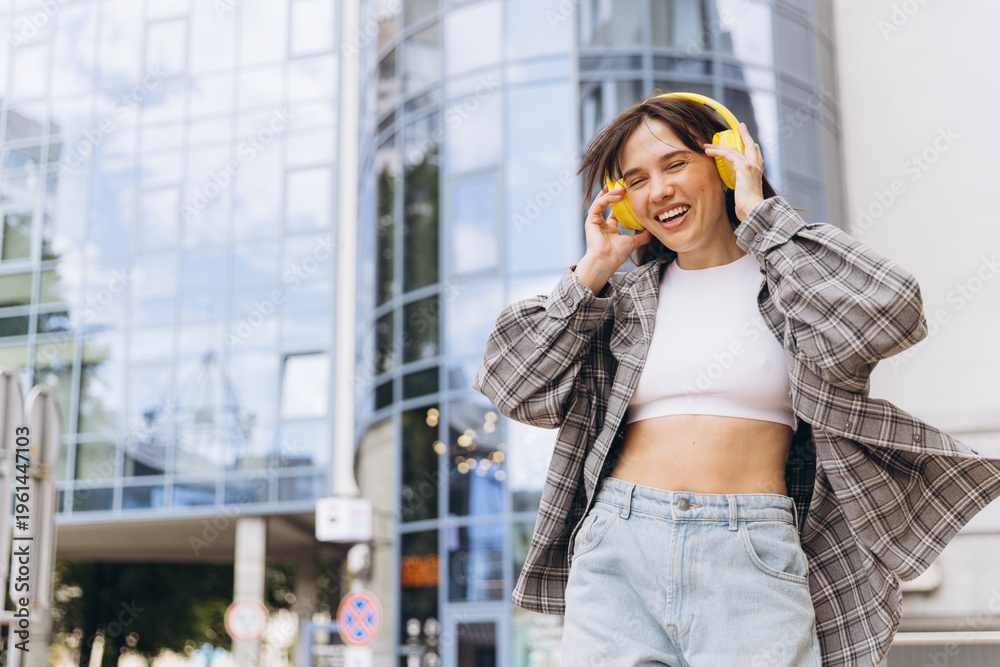 Fototapeta premium Young woman enjoying music with yellow headphones, smiling happily in city street, urban lifestyle