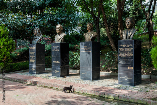 A stray cat strolls past bronze busts of world historical figures in a leafy Istanbul park garden, Kadikoy