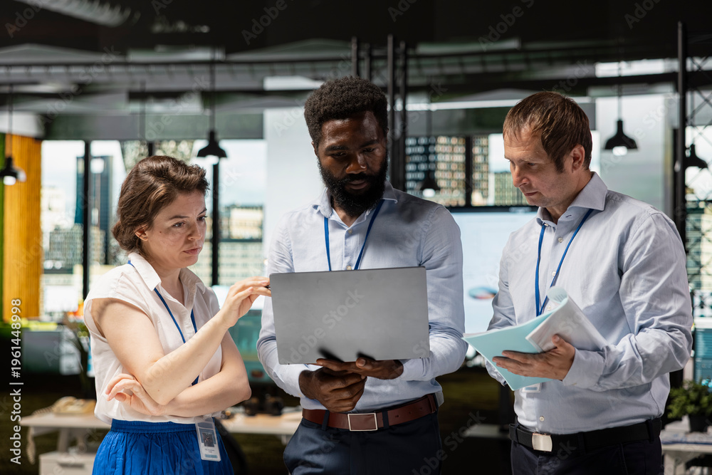 Fototapeta premium Black man and two white colleagues working together on financial reports and business investment strategies. Multiethnic office team standing and reviewing company performance on laptop.