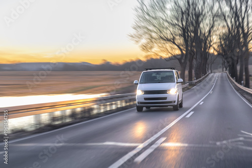 Quadro su tela White minivan driving on rural road near river at sunset