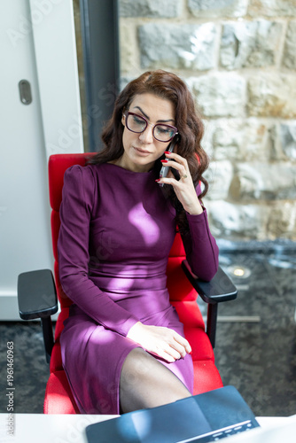 Caucasian woman winding down after work call seated in red chair near stone wall wearing purple dress and glasses hands resting on lap desk with soft sunlight contemplative mood and gentle