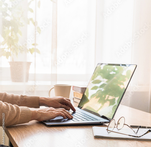 Womens hands typing on laptop keyboard, working remotely from home office desk with mug and glasses. Person typing on laptop working from home