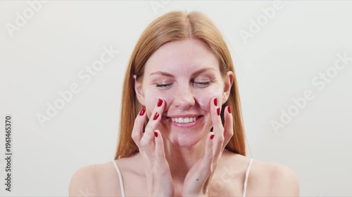 Red-haired Woman smiles and touches her face while showing her red nail polish in a bright indoor setting during the day