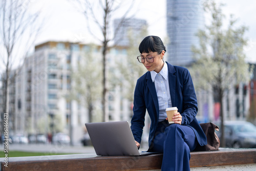 Fotografie Businesswoman working on laptop outdoors with coffee, urban background