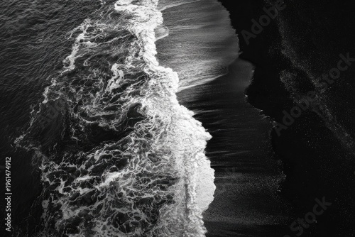 Aerial, monochrome view of ocean waves crashing onto dark sand. Contrast between foam and shore