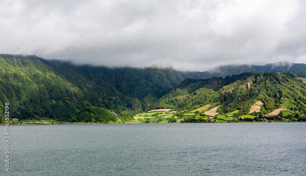 Fototapeta premium Crater lake at Sete Cidades, San Miguel, Azores. Volcanic caldera lake surrounded by lush green hills. Cloudy sky over serene water landscape. Located in Portugal.