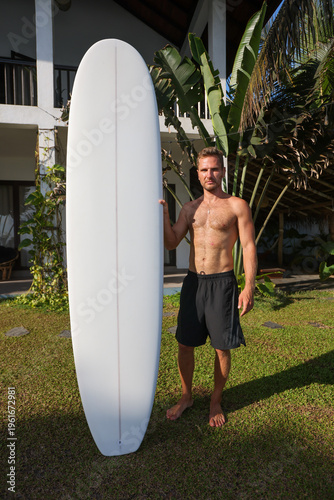 Confident Surfer In Casual Attire Poses Beside Palm Trees With Lengthy Board In Tropical Setting