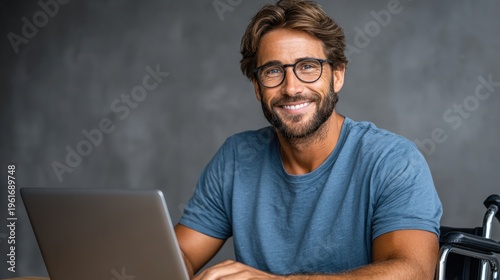 Smiling positive young adult in wheelchair using laptop with glasses, wearing bright blue t-shirt against gray backdrop, expressing happiness and focus, technology, inclusivity, accessibility