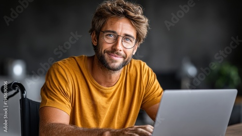 Smiling positive young adult in wheelchair using laptop with glasses, wearing bright yellow t-shirt against gray backdrop, expressing happiness and focus, technology, inclusivity, accessibility