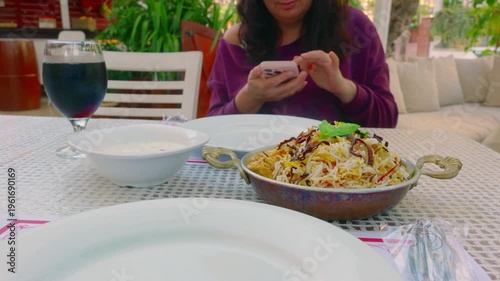 Person checks device prior to meal at cafe. Female individual views smartphone while seated at dining table. Woman pauses to examine her phone as she relaxes at cafe patio
