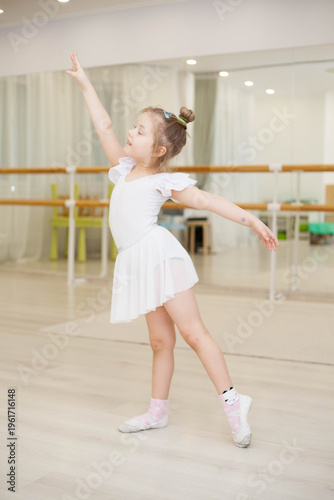 Fotografie A little girl in a white bodysuit practices ballet in a mirrored studio, trainin