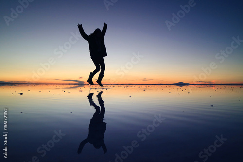 Silhouette of a happy traveler jumping on the amazing mirror effect of Uyuni Salt Flats at sunset, Bolivia, South America