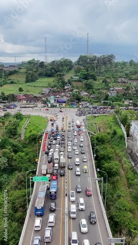 Aerial view of heavy traffic congestion on a bridge near a rural town, with long lines of vehicles under cloudy skies surrounded by green hills and scattered houses