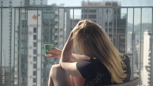 Woman sits on balcony in city with coffee cup while looking at skyline on sunny day in afternoon