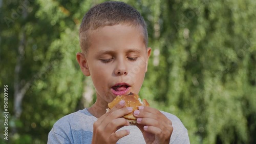 Cute 9-year-old boy eating a burger in a park. Concept of children's nutrition, fast food, outdoor snack, and lifestyle. Childhood, leisure, and eating habits in an urban green space.