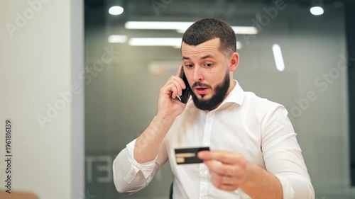 Businessperson engaged in phone conversation, reviewing credit card. Displays focused attention and potential concern during a financial transaction. Modern communication and digital banking concepts.