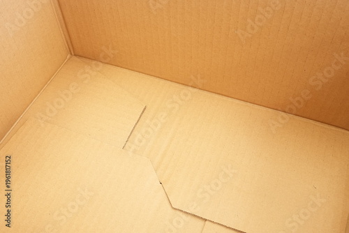 Empty cardboard box rests quietly inside a corner. Wrinkles and creases tell stories of past use. Smooth interior shows gentle, natural brown tones. Simple, uncluttered space invites imagination