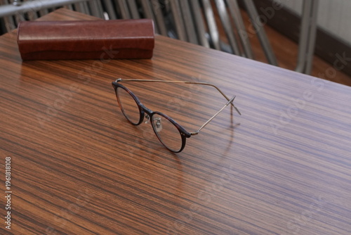 Round glasses rest on a polished wood table. Brown case lies nearby, softly shadowed. Sleek frame reflects subtle light. Stillness speaks of quiet thought or pause