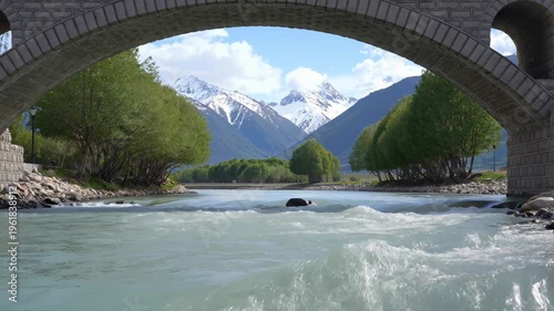 Scenic View of a Rushing River Under a Stone Bridge with Snow-Capped Mountains in the Background. Seamless loooing 4k video background