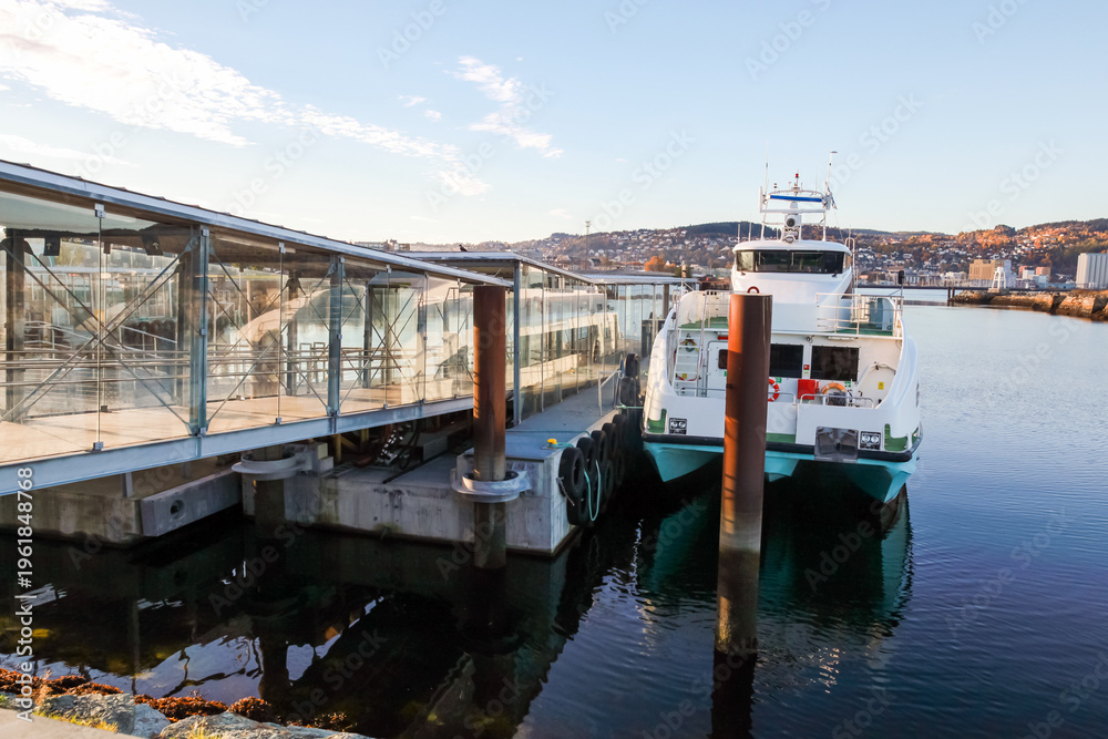 Fototapeta premium Trondheim fast boat terminal coastal view. A modern ferry moored at a glass-sided terminal along a calm waterfront, city skyline and hills in the background. Norway