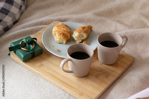 Bamboo serving board holding two ribbed coffee mugs and croissant halves on bed with check pillow