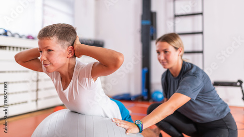 Boy performing a motoric corrective core exercise on a stability ball while a trainer supports trunk positioning and supervises controlled movement during functional strength development.