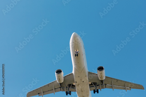 Bottom view of a passenger airplane flying overhead with landing gear extended against a clear blue sky.