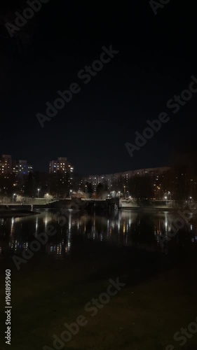 City lights reflecting in calm lake at night, quiet residential waterfront scene with apartment buildings, urban park atmosphere and moody evening skyline background.