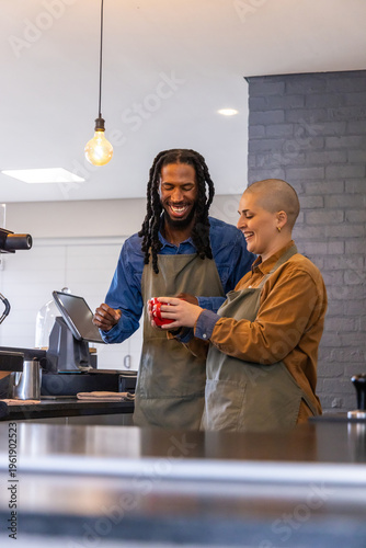 Diverse coworkers smiling and making coffee at cafe counter, wearing aprons, handling red cup