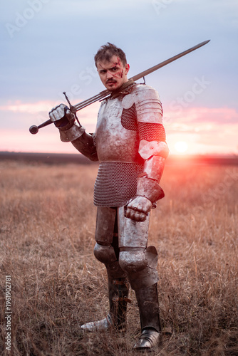 Photos A medieval knight with a bloody face, dressed in plate armor with a sword and a torch in his hands, poses standing in a field against the backdrop of the sky and sunset