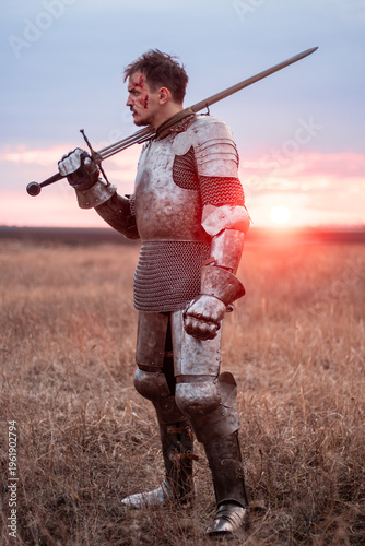 Tableau sur toile A medieval knight with a bloody face, dressed in plate armor with a sword and a torch in his hands, poses standing in a field against the backdrop of the sky and sunset
