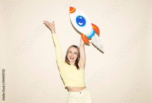 Smiling woman in yellow outfit holding handmade paper rocket celebrating success