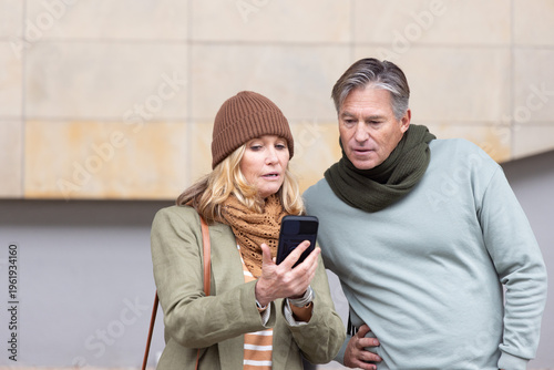 Senior couple studying smartphone while standing in front of beige tiled facade, holding bag
