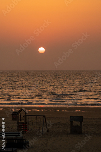 A stunning sunset casts a warm hue over the calm ocean, creating a peaceful ambiance at the beach, complemented by the silhouette of playground equipment in the foreground.