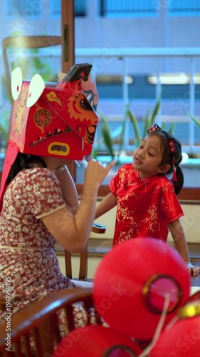 Thai mother and young daughter in traditional red Qipao playing with DIY Chinese dragon mask during Lunar New Year celebration at home.