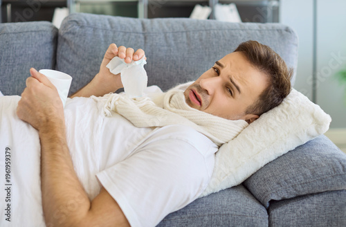 Sick man with stuffy runny nose, allergy or viral fever resting on sofa at home. Sad person in scarf holding tissue and mug, holding mug of hot drink, suffering from fatigue, headache and inflammation