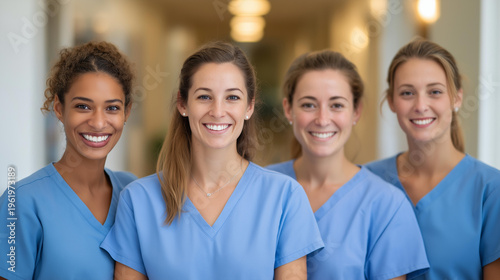 Group portrait of young nurses standing together in a bright hospital hallway, confident smiles, matching scrubs, defocused medical carts behind them, nursing team, healthcare workforce,