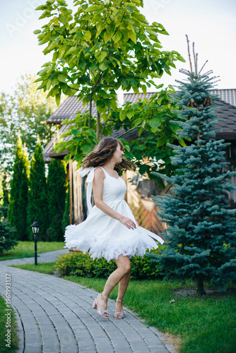 Happy bride in short white wedding dress walking along garden path outdoors. Soft natural light, greenery and elegant details create romantic atmosphere, capturing joyful moment of wedding day.