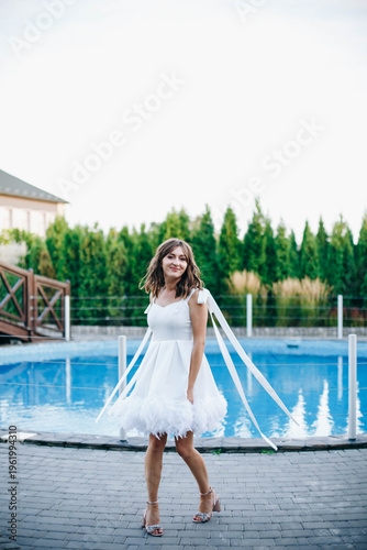 Young woman in white dress with feather hem posing near swimming pool, stylish summer portrait. Elegant feminine look, natural light, modern outdoor lifestyle scene with water reflections.