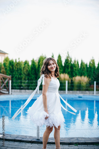 Young woman in white dress with feather hem posing near swimming pool, stylish summer portrait. Elegant feminine look, natural light, modern outdoor lifestyle scene with water reflections.
