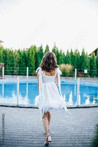 Young woman in white dress with feather hem posing near swimming pool, stylish summer portrait. Elegant feminine look, natural light, modern outdoor lifestyle scene with water reflections.